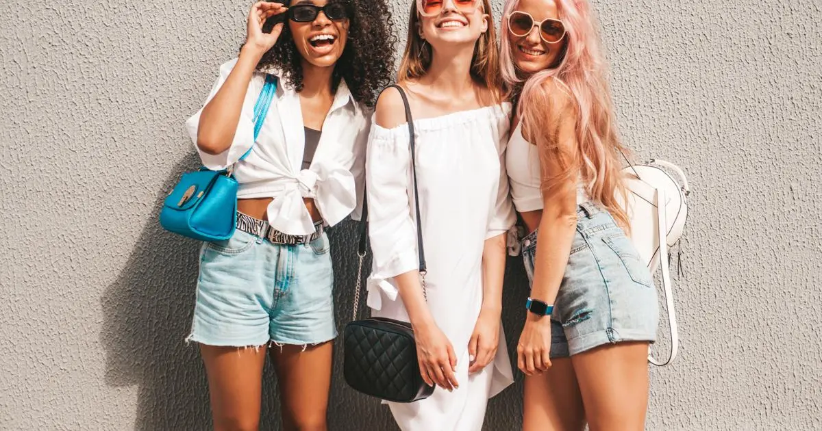Três mulheres de óculos escuros e shorts posando para uma foto, irradiando confiança e aproveitando um dia ensolarado.